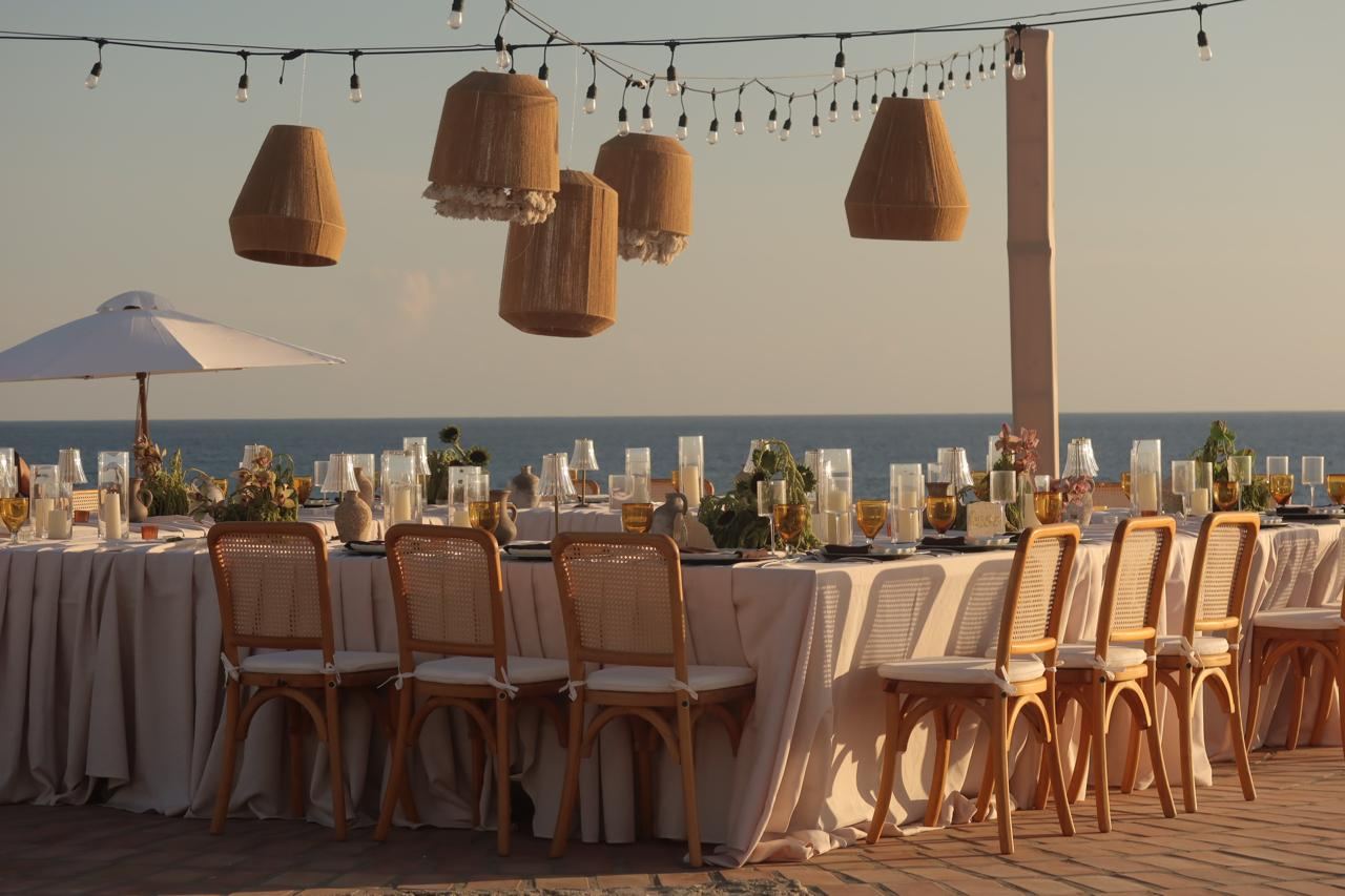 Long dinner table with woven lanterns at golden hour, ocean behind
